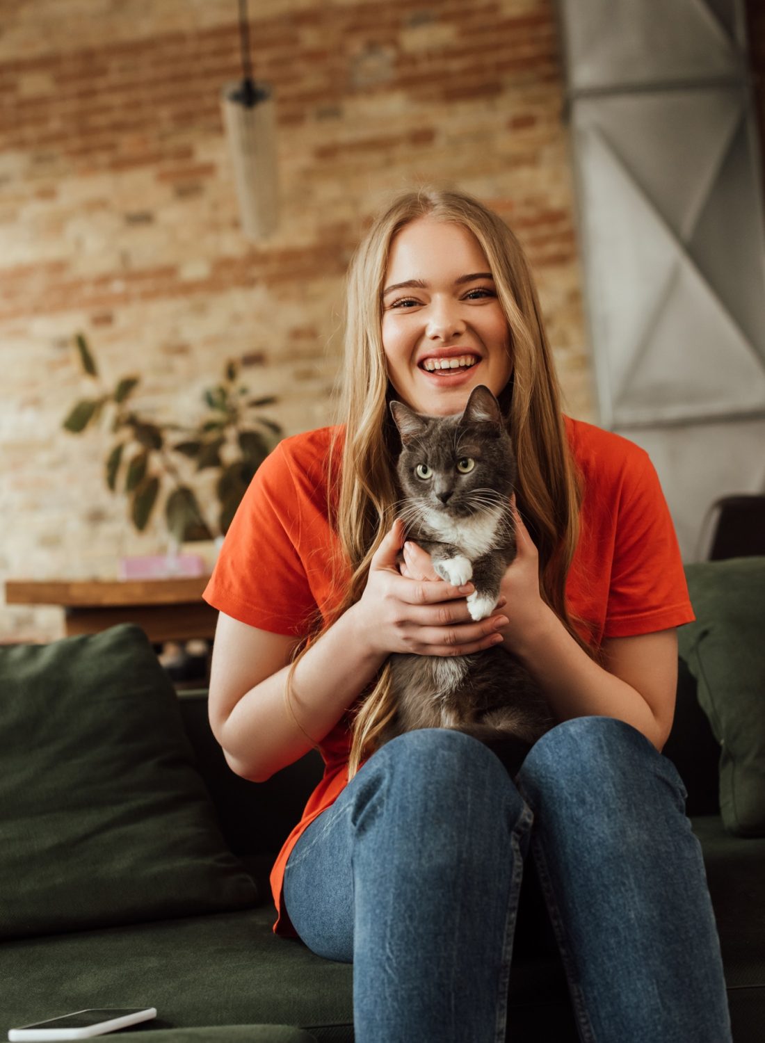 happy young woman holding in arms cute cat near smartphone on sofa